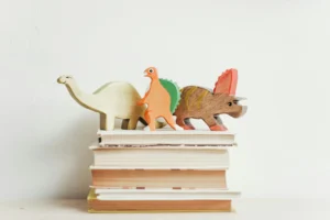 There is a stack of about five books sitting with their spines faced away from the viewer on a white desk and in front of a white wall. Only the edges of their pages are visible, and these give no clues about their contents.<br /> On top of the books are three toy wooden dinosaurs: a white brontosaurus, a brown stegosaurus with red horns, and a third orange dinosaur with a green spine on its back whose proper name I can’t identify. It is standing on its hind legs.