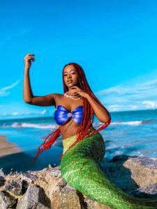 A gorgeous black woman dressed in a mermaid costume and sitting on the sand at the beach as the waves roll in. Her bra is metallic purple, her fin is light green, and her hair has been braided with red extensions. She’s lovely and looks very happy to be there. 