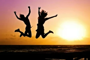 The silhouttes of two people with long straight hair who are leaping for joy on the beach as the sun sets. The ocean water behind them is rippling gently. 