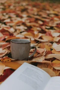 Photo of a light brown mug and an opened book lying in a bed of red and yellow autumn leaves on the forest floor. 