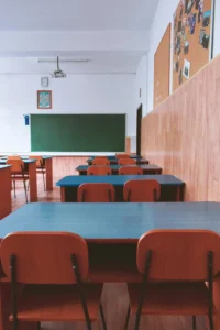 Photo of rows of empty, two-person desks in a classroom. Each desk has two chairs and enough space for two kids to sit there. You can see a chalkboard at the front of the room but it has been wiped clean and is ready for the next class.