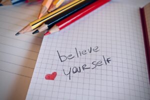 The phrase “believe yourself” written in black coloured pencil on white graph paper. There’s a little red heart written in red coloured pencil next to that phrase as well as three coloured pencils - gold, black, and red - lying on the page above these scribbling. 