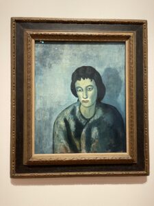 A photo of a painting called “Woman With Bangs” by Pablo Picasso. The painting is of a sad-looking woman who does, indeed, have bangs in her dark brown hair. She is looking morosely to the side in this painting. There is a blue hue to the painting that only makes it look even sadder. 