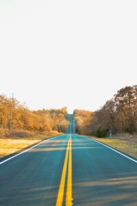 A photo taken of an empty country road in late autumn. All of the trees on both sides of the road have revealed their fall colours, and some of the trees are beginning to look sparse as their yellow, red, orange, or brown leaves fall off. 