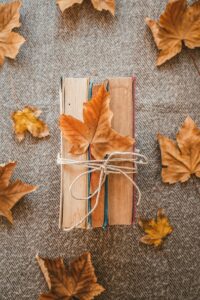 A photo of three hardcover books bound together tightly by a thick piece of twine and placed on their spines. There is an orange leaf tucked into the twine just below the knot in it as well as some other orange and brown autumn leaves scattered on the burlap cloth these books are sitting on. 