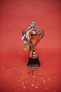 A photo of a gold-coloured championship cup with two handles. It’s the sort of award that would typically be given to someone who won first place. There are two gold-coloured medallions with red, white, and blue ribbons on them hanging from the cup. There is also gold-coloured glitter on the red surface where this cup is sitting. It looks like someone just had a celebration here! 