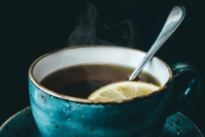 Closeup photo of a teal teacup filled with steaming hot tea. The inside of the cup is white which I think is a poor choice for such a staining liquid. There is a thin slice of lemon floating in the tea and a spoon resting in the tea as well. A little sugar must have just been mixed in. You can see steam lifting from the tea, so it still must be pretty hot! Behind the teacup it is dark and not much can be seen other than the gentle little handle of the cup that curves gracefully out so that one or two fingers can lift it. 