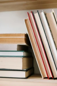 A stack of five books sitting on a shelf with their spines to the left out of view of the viewer. A second stack of five slimmer volumes are leaned up against the first stack, and these, too, have spines we can’t read. Their spines are turned toward the white wall. 