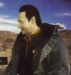 Brent Spiner playing Data on the set of "Star Trek: Nemesis.” He’s wearing a black jacket and looking off to the side as someone out-of-view speaks to him. In the background there is a set that shows a blue sky with a few wispy white clouds in it and a hilly, desert landscape behind him. Data looks like a very pale white man with black hair, if you’re not aware of what this character looks like. He’s an android but otherwise looks like a human when his complexion isn’t taken into account. 