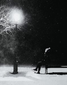 Photo of a white woman sitting on a bench on a cold wintry night after dark. She is wearing a winter hat and warm coat and staring up at a streetlamp next to the bench. The light from the lamp illuminates the surroundings just enough for the viewer to see some snow gently falling down to the ground. This is a cold scene due to the weather, but the woman looks contemplative as she gazes upon the street lamp. I’d sit by her. 