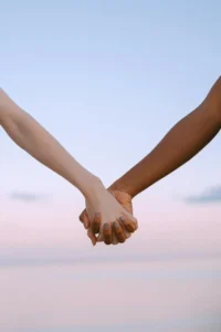 The bare arms of two people who are holding hands. The background shows a pink sky that hints these people are watching either a sunset or a sunrise. One of the people holding hands has brown skin while the other person has pale skin. 