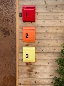 Photo of three small mailboxes that have been installed on a wooden walls. The mailboxes are numbered 1 through 3 and are painted red, orange, and yellow respectively. You can also see a few branches from a bush growing in the lower right corner of the photo. 