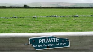 Photo of a wooden fence with barbed wire strung at the top of the fence. The sign on the fence reads, “private no public right of way.” On the other side of the fence you can see a large grassy area and, in the distance, a grove of trees separating this grazing area from the next one. 