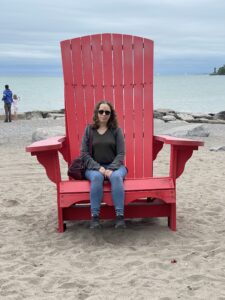 Lydia, a white woman with curly brown hair who is wearing jeans, sneakers, and a light jacket, is sitting in a comically oversized red beach chair. She is smiling slightly. 