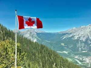 A Canadian flag flying on a flagpole in front of the Banff National Park mountains which are large and spectacular. There’s a large hill or maybe a small mountain between the flagpole and Banff that’s covered in fir trees. 
