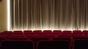 Photo of the red, soft chairs in an upscale movie theater. There is a curtain covering the screen and light glowing at the bottom of the curtain, probably to give audience members a better chance to see where they’re walking before the film begins. 