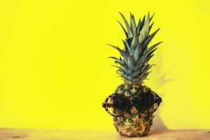 A pineapple sitting in front of a bright yellow wall. It has a pair of sunglasses hanging on the fruit as if the pineapple has eyes. The green leaves of the pineapple are still attached to it and look like a spiky hairdo. 