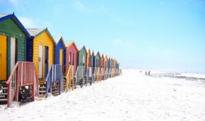 A row of colourful beach huts painted red, blue, yellow, green, and other primary colours. The beach has white sand and looks pristine. 