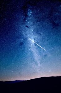 A shooting star falling in front of the Milky Way just after dusk. The land below is treeless and rather flat, so the sky is the star of this show. The sky moves from being dark blue to light blue to showing just a sliver of light from the sun that’s just set over the horizon as you move your eyes from the top of the scene to the bottom of it. 