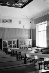 Black and white photo of a vintage classroom where each wooden desk sits two pupils. The room is empty and feels a little desolate. 