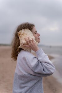 A white woman wearing a grey sweatshirt is listening to a conch shell at the beach on an overcast and what appears to be somewhat chilly day. 