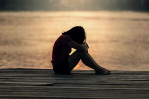A person with long straight hair is sitting on a pier at sunset and leaning their head on their knees. Their body language looks bent over and sad. 