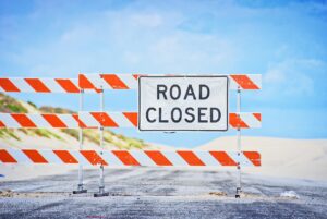 Photo of a road closed sign that has been erected in the middle of a road that looks like it’s headed toward the beach. 