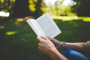 A pale-skinned person wearing jeans is sitting in the park and reading a book as the sunlight streams down onto the grass and trees. 
