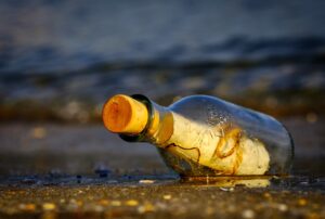 A glass bottle that has a piece of paper sealed inside of it. The bottle is sitting in the sand at the beach and you can see the water coming in behind it. No idea what message is scrawled on the paper, though!