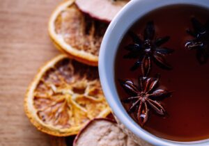 Star anise soaking in some dark tea in a white teacup. Beside the teacup on a wooden table are two slices of dehydrated oranges. 