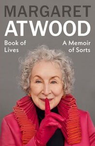 Book cover for Book of Lives by Margaret Atwood. Image on cover shows Ms. Atwood smiling and holding one finger up to her lips as if to shush the viewer. She’s wearing a bright read sweater and red gloves, too. 