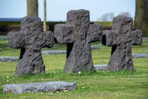 Three grey and roughly hewn stone crosses in a German military cemetery from World War I. The sky is blue and the grass is green. It’s a peaceful scene. 