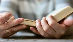 Closeup photo of someone’s hands as they hold open a book with two hands and read it. They’re wearing a wedding ring and have pale skin. Their age and sex isn’t easy to determine. 