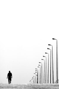 Black and white photo of someone walking down a desolate road next to a line of street lamps that are closely packed together but not turned on.