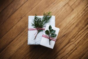 Two gifts, one the size of a hardback novel and the other the size of a large adult human hand, are wrapped in white paper. They both have red ribbons tied around them and greenery tucked inside of the ribbon next to the wrapping paper. 