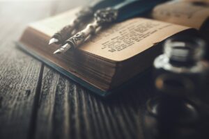 Two ballpoint pens lying on an opened hardcover book that’s filled with beautiful calligraphy. Nearby is an inkwell. 