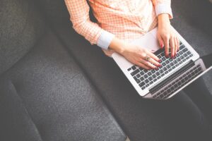 A white person wearing a light orange, long-sleeved shirt is typing on a laptop while sitting on a couch. 