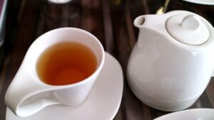 A white teacup filled with plain tea. Beside it is a white tea maker. 