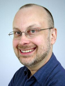 Photo of famous Canadian science fiction author Robert J. Sawyer, a middle-aged white man wearing a dark blue button-down shirt and glasses. His hair is cut short and he’s smiling broadly in this shot. 