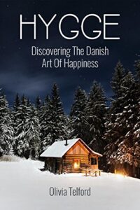 Book cover for Hygge - Discovering the Danish Art of Happiness by Olivia Telford. Image on cover shows a small log cabin with lamplight glowing out of its window. There’s a thick layer of snow outside, the sky overhead is turning from dusk to dark, and there is a pine forest behind the cabin. 