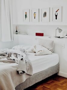 A photo of an unmade bed whose white rumpled covers have been pulled back to air out the white sheets for a while. Or maybe this person simply didn’t want to make the bed! Above the bed there is a headboard that has a small lamp on it as well as a series of paintings of four colourful and possibly tropical birds hanging on the white wall above. 