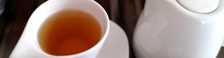 A white teacup filled with plain tea. Beside it is a white tea maker.